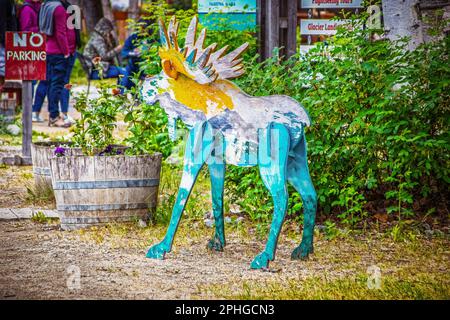 Alaska tourism - Roughly painted board moose standing by tourist signs for businesses and wooden flower planters - unrecognizable people lined up in b Stock Photo