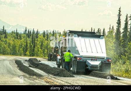 Roadwork on blacktop highway with workman in safety shirt by truck with dust and smoke  spitting out rows of Bituminous asphalt - pine forests and mou Stock Photo