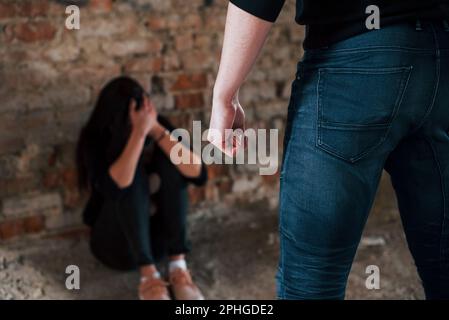 Violent man standing and threatens girl that sits on the floor with teddy bear in abandoned building Stock Photo