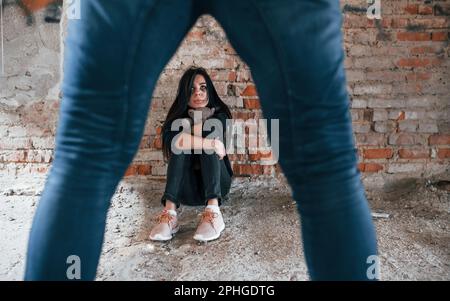 Violent man standing and threatens girl that sits on the floor with teddy bear in abandoned building Stock Photo