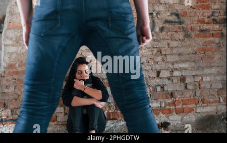 Violent man standing and threatens girl that sits on the floor with teddy bear in abandoned building Stock Photo