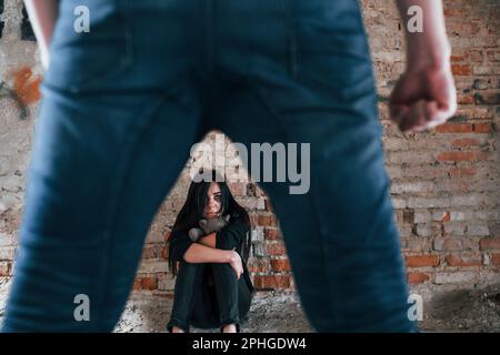 Violent man standing and threatens girl that sits on the floor with teddy bear in abandoned building Stock Photo