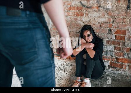 Violent man standing and threatens girl that sits on the floor with teddy bear in abandoned building Stock Photo