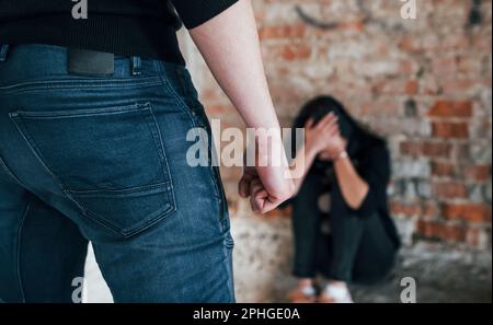 Violent man standing and threatens girl that sits on the floor with teddy bear in abandoned building Stock Photo