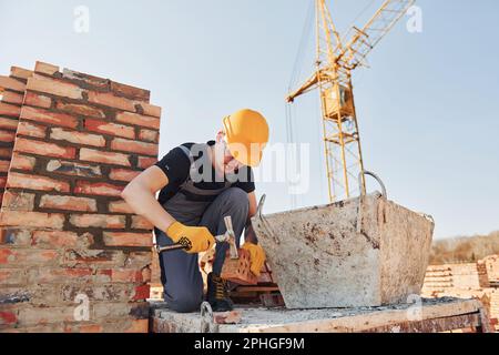 Holding brick and using hammer. Construction worker in uniform and safety equipment have job on building Stock Photo
