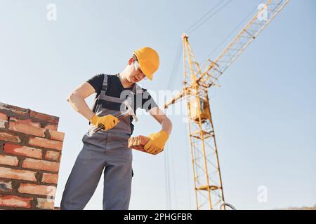 Holding brick and using hammer. Construction worker in uniform and safety equipment have job on building Stock Photo