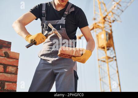 Holding brick and using hammer. Construction worker in uniform and safety equipment have job on building Stock Photo