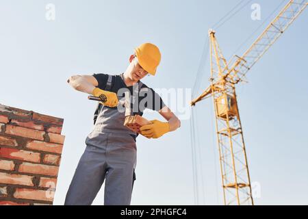 Holding brick and using hammer. Construction worker in uniform and safety equipment have job on building Stock Photo