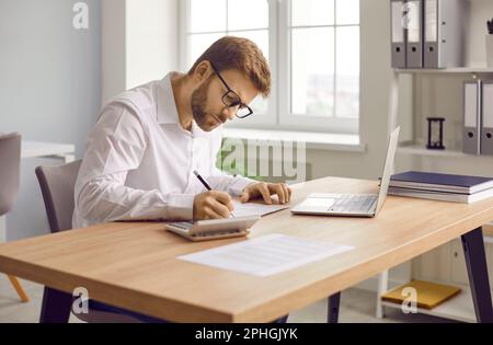 Focused businessman sitting at desk using laptop and making notes on paper Stock Photo