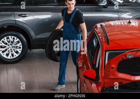 Worker in black and blue uniform holds car wheel and walks with in near red automobile Stock Photo