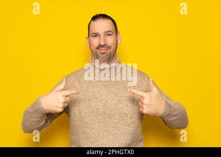 The portrait of a bearded handsome man points to himself puzzled at being chosen. Amazed and attractive hispanic man over yellow background, he has a Stock Photo