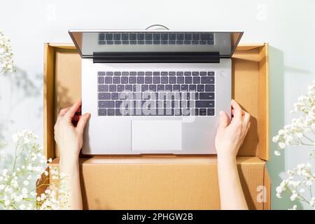 Buying and delivering new laptop. Romantic overhead shot of womans hands holding laptop in cardboard box on blue background with white spring flowers Stock Photo