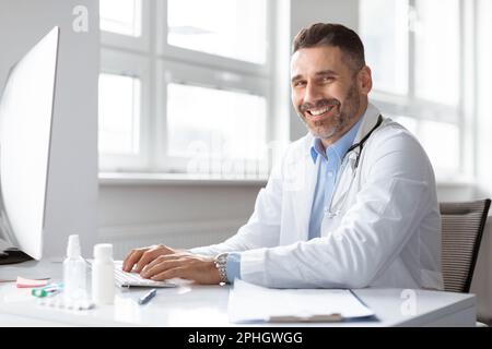 Happy male doctor medic expert using computer at work in hospital, checking patients electronic files medical forms Stock Photo