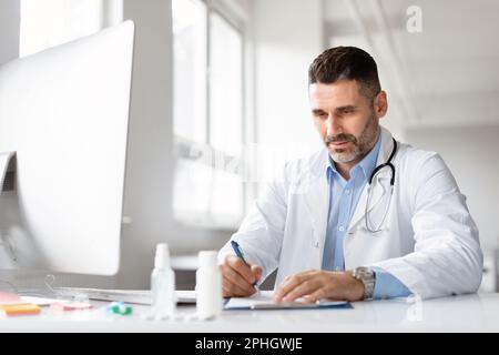 Serious male doctor using computer and writing notes in medical journal sitting at workplace in clinic Stock Photo