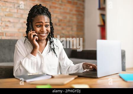 Mobile communication. Black woman talking on cellphone working on laptop at home, having phone conversation Stock Photo