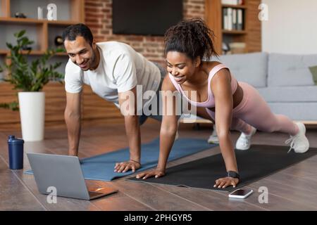 Smiling young black couple in sportswear watch video on laptop, doing push-ups on floor, workout together Stock Photo