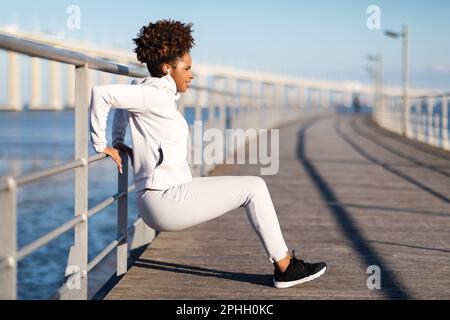 Young black woman in sportswear squatting outdoors, using handrail as support Stock Photo