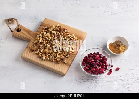 Wooden board with chopped walnuts, bowls with dried cranberries and spices on a light gray textured background, top view. Ingredients for baked biscot Stock Photo