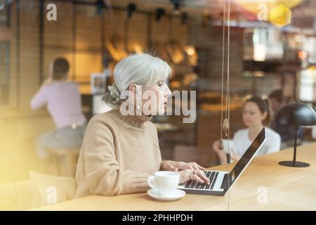 Aged female freelancer sitting with coffee and laptop in cafe Stock Photo