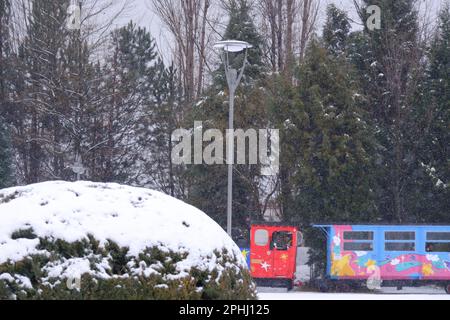 Colorful train for children in a snowy winter day. Train moving behind trees and bushes seen at distance. Stock Photo