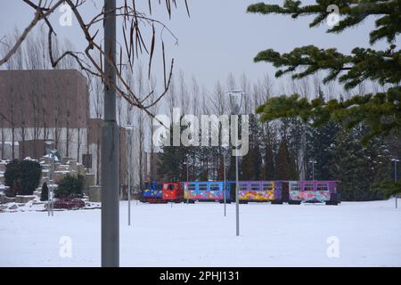 Colorful train for children in a snowy winter day. Train moving behind trees and bushes seen at distance. Stock Photo