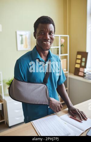 Vertical portrait of black young man with arm sling working at standing desk and smiling at camera Stock Photo