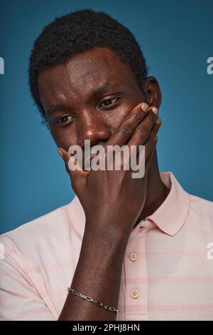 Vertical closeup portrait of distressed black man covering mouth against blue background Stock Photo