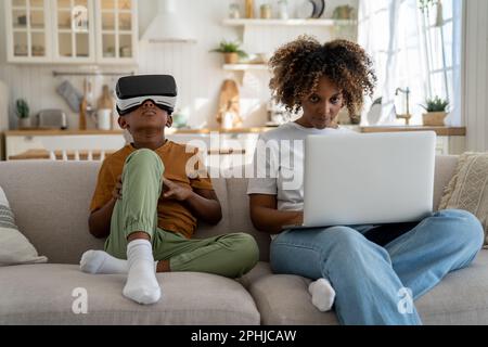 Child using virtual reality simulator headset while sitting on sofa with mother working on laptop Stock Photo