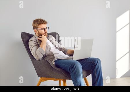 Young programmer is sitting in armchair, working on laptop computer, and thinking Stock Photo