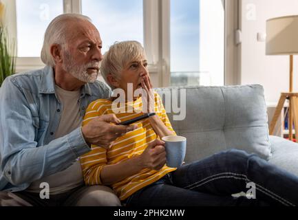 Senior couple watching scary scenes in movie on sofa at home. Mature man holding remote control Stock Photo