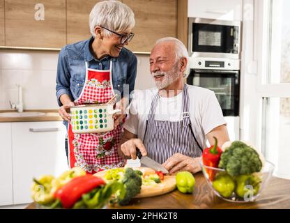 Cheerful senior couple preparing soup, husband cutting vegetable while wife holding pot and smiling Stock Photo
