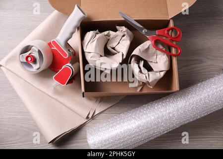 Open box with wrapped items, adhesive tape, scissors, paper and bubble wrap on wooden table, flat lay Stock Photo