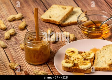 Peanut butter or peanut paste in an open jar and spread on a toast sandwich with banana and honey and peanuts in shells scattered on the table Stock Photo