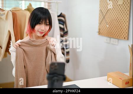 Charming young Asian female entrepreneur clothes shop owner live streaming through online shopping platform. SME business concept Stock Photo