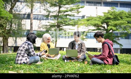 A group of happy and smart young Asian-diverse college students are using laptop and working on their school project in the campus park together. Uni- Stock Photo