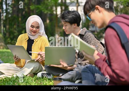 Attractive and friendly young Asian Muslim female college student talking and discussing work with her male friend while sitting on the grass in the c Stock Photo