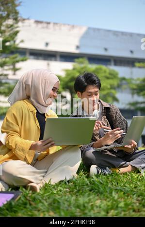 Happy young Asian Muslim female college student sharing her ideas and discussing work with her male friend while relaxing in the campus park together. Stock Photo