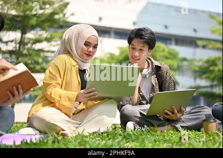 Focused young Asian Muslim female college student discussing work with her male friend while relaxing in the campus park together. Stock Photo
