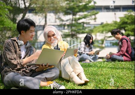Two Asian college students are talking and working on their schoolwork while sitting on the grass and relaxing in the campus park together. Stock Photo