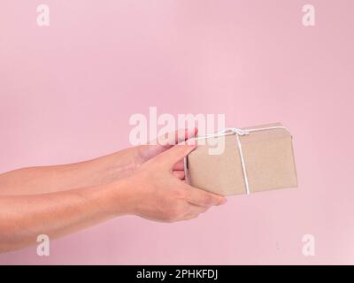 Close-up of female hand holding present box or gift box in craft paper on pink background. Small gift in female hands indoor. Stock Photo