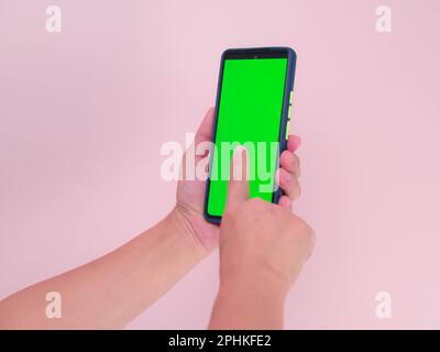 Human hand using mobile smartphone with green screen isolated on pink background. Woman hand scrolling feed on smartphone with green screen mockup. cl Stock Photo