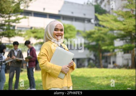 Attractive and smiling young Asian Muslim female college student in hijab carrying her portable laptop and standing in the campus park. Stock Photo