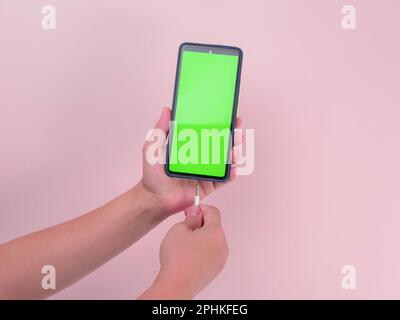 Female hands plugging earphone into smart phone with green screen isolated on pink background. Close-up of hand plugging in headphone device in smartp Stock Photo