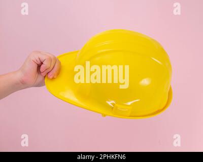 Close-up of female engineer hand holding yellow safety helmet on pink background. Human hand holding a hard hat. Stock Photo