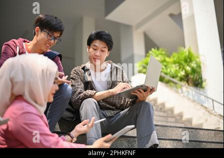 A group of Asian-diverse college students are talking, relaxing and working on their schoolwork on the stairs outside of the campus building together. Stock Photo
