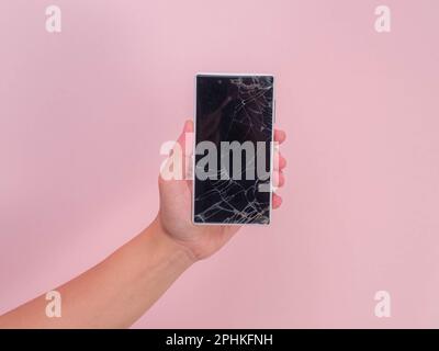 Close-up of hand holding mobile phone with broken touch screen on pink background. Female hand holding old phone with cracked and damaged screen. Stock Photo