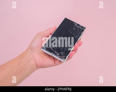 Close-up of hand holding mobile phone with broken touch screen on pink background. Female hand holding old phone with cracked and damaged screen. Stock Photo