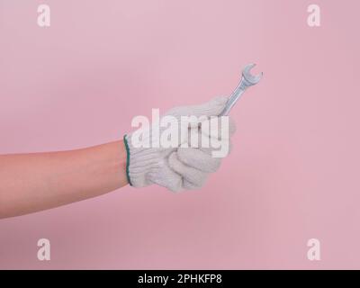 Hand in white knitted glove holding metal wrench isolated on pink background. Worker's hand in a glove holding a wrench. Stock Photo