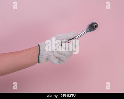 Close-up of hand in white knitted glove holding socket wrench isolated on pink background. Hand in protective glove holding a ratchet wrench (socket). Stock Photo