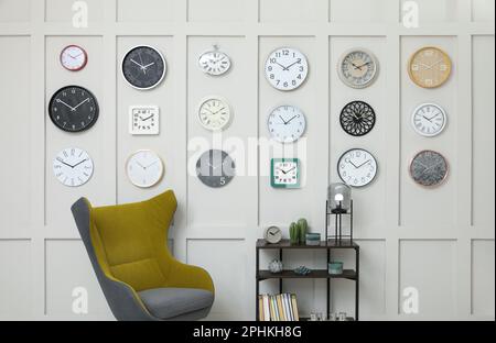 Armchair, console table and many different clocks hanging on white wall in room Stock Photo